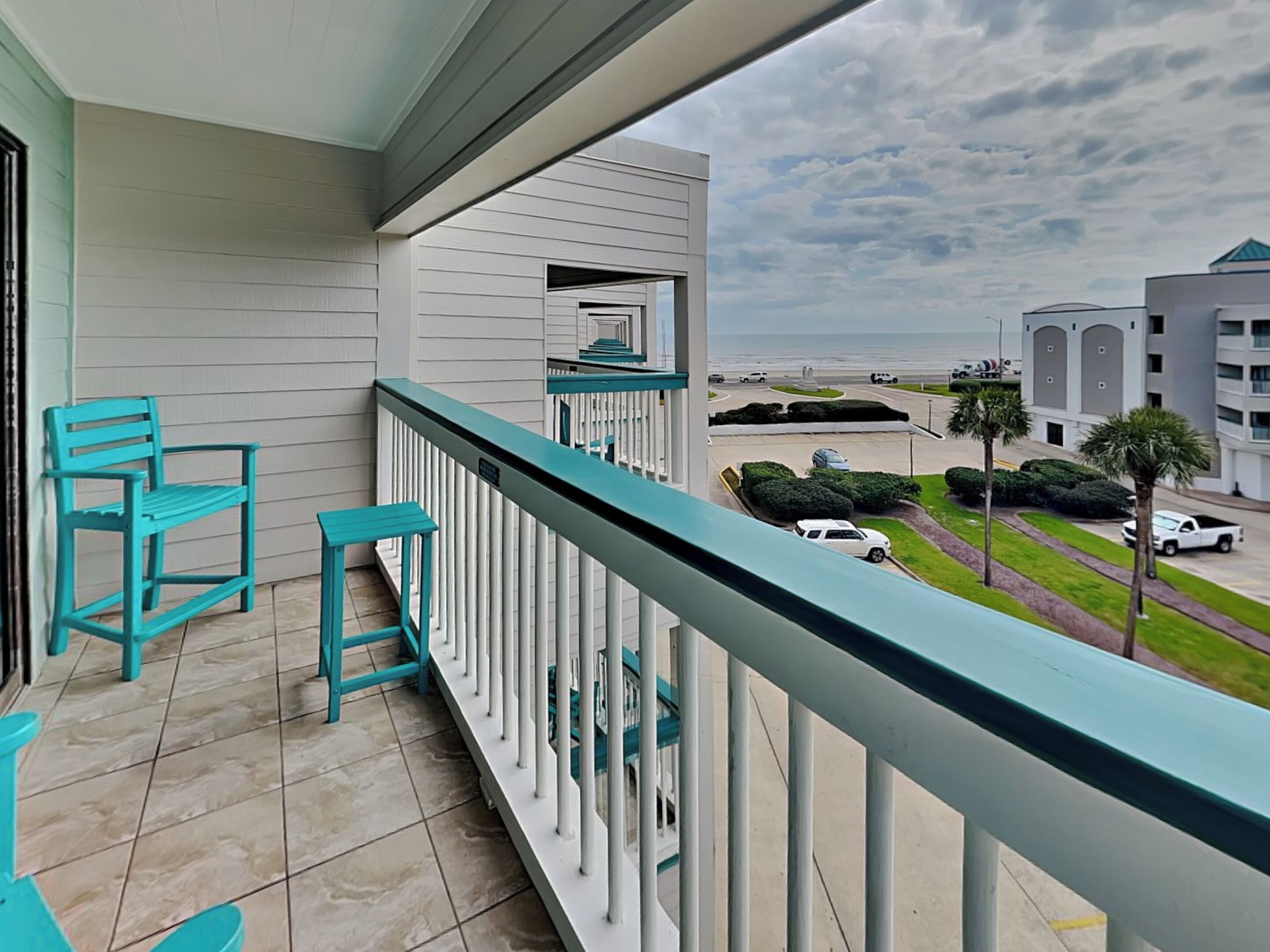 A balcony with teal chairs and a small table, overlooking a parking area and buildings.