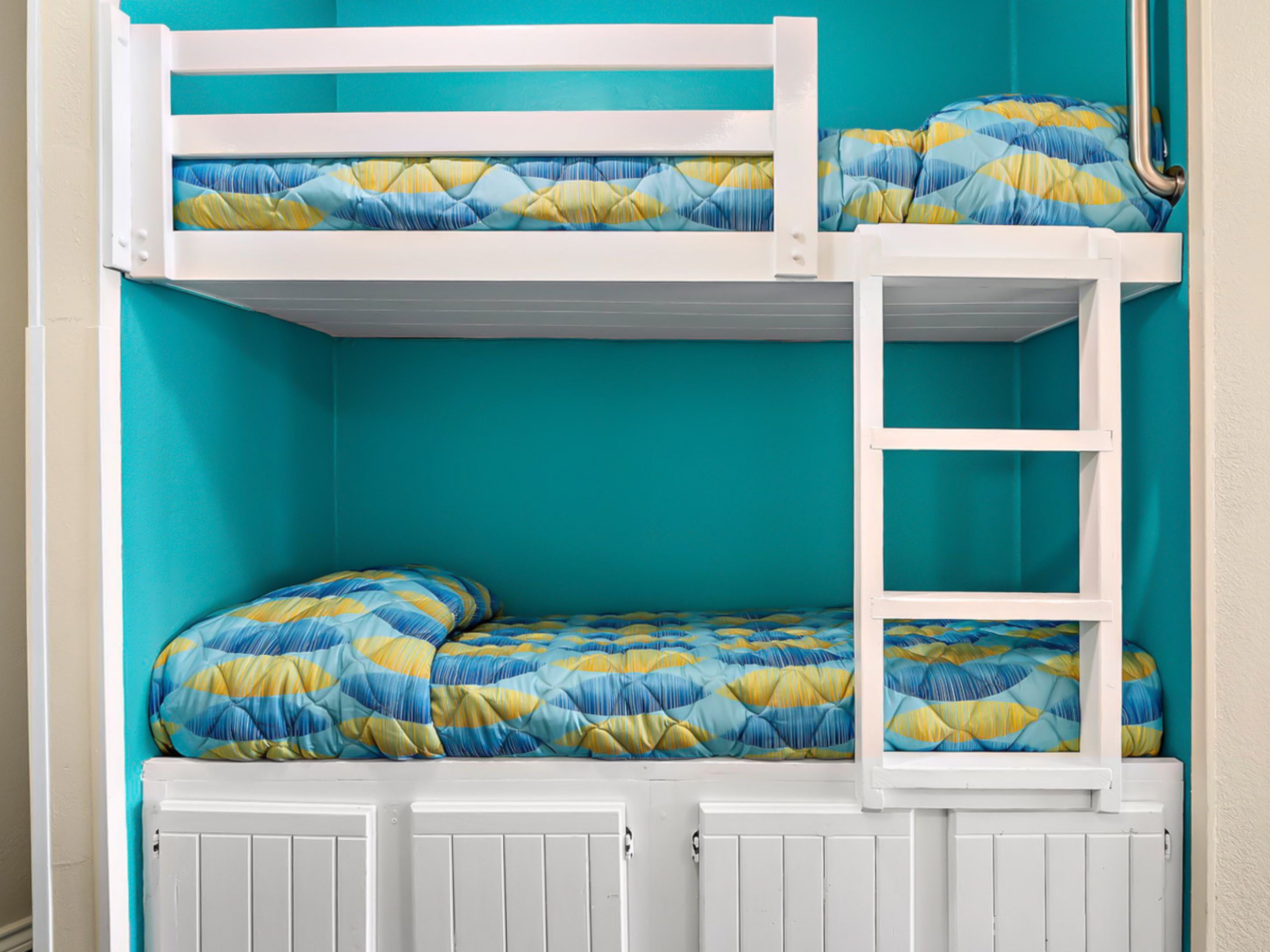 A bunk bed with a blue and yellow patterned bedspread, white railings, and a white door below.