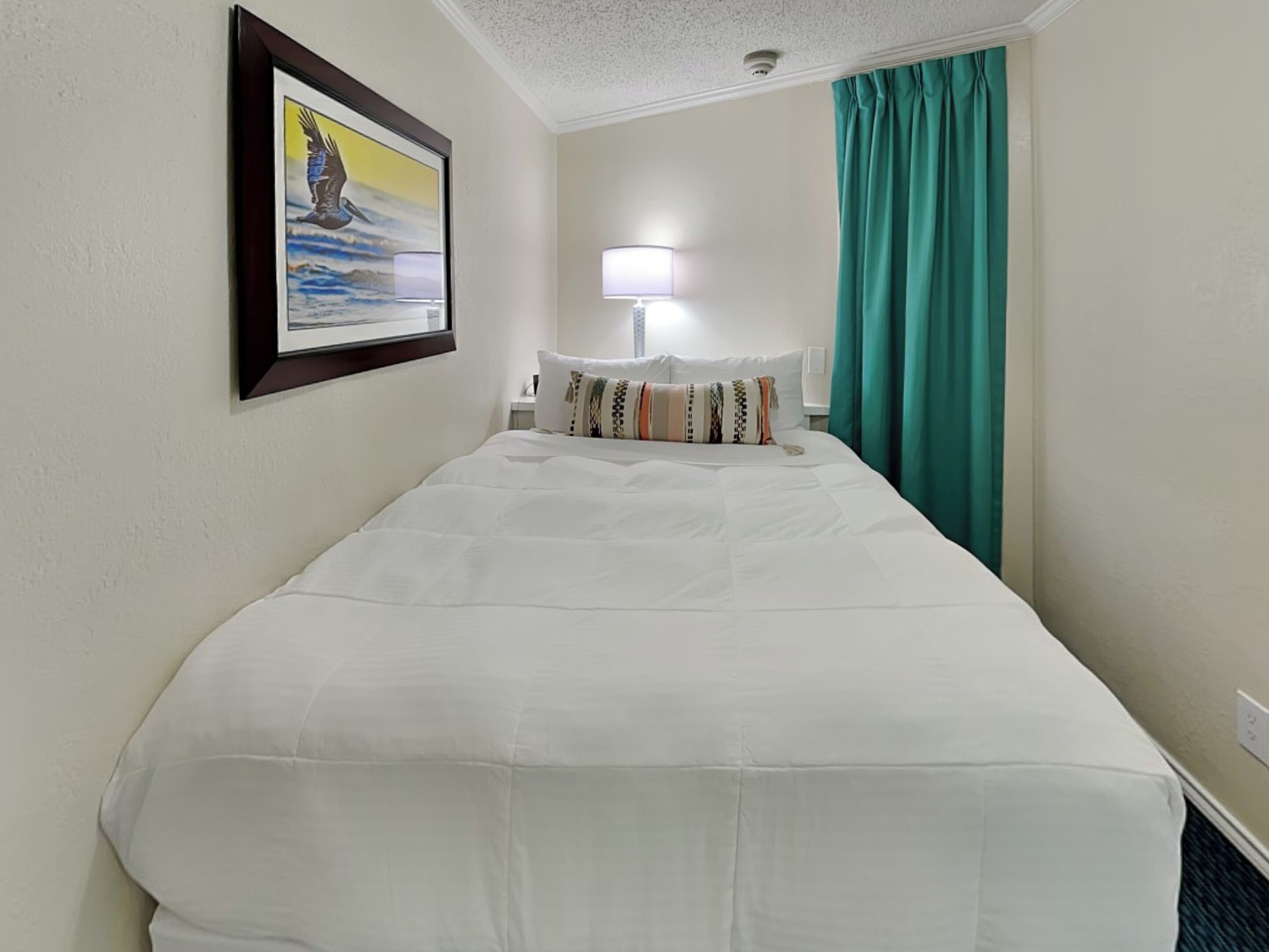 A hotel room with a large bed, a nightstand, a lamp, a painting on the wall, and teal curtains.