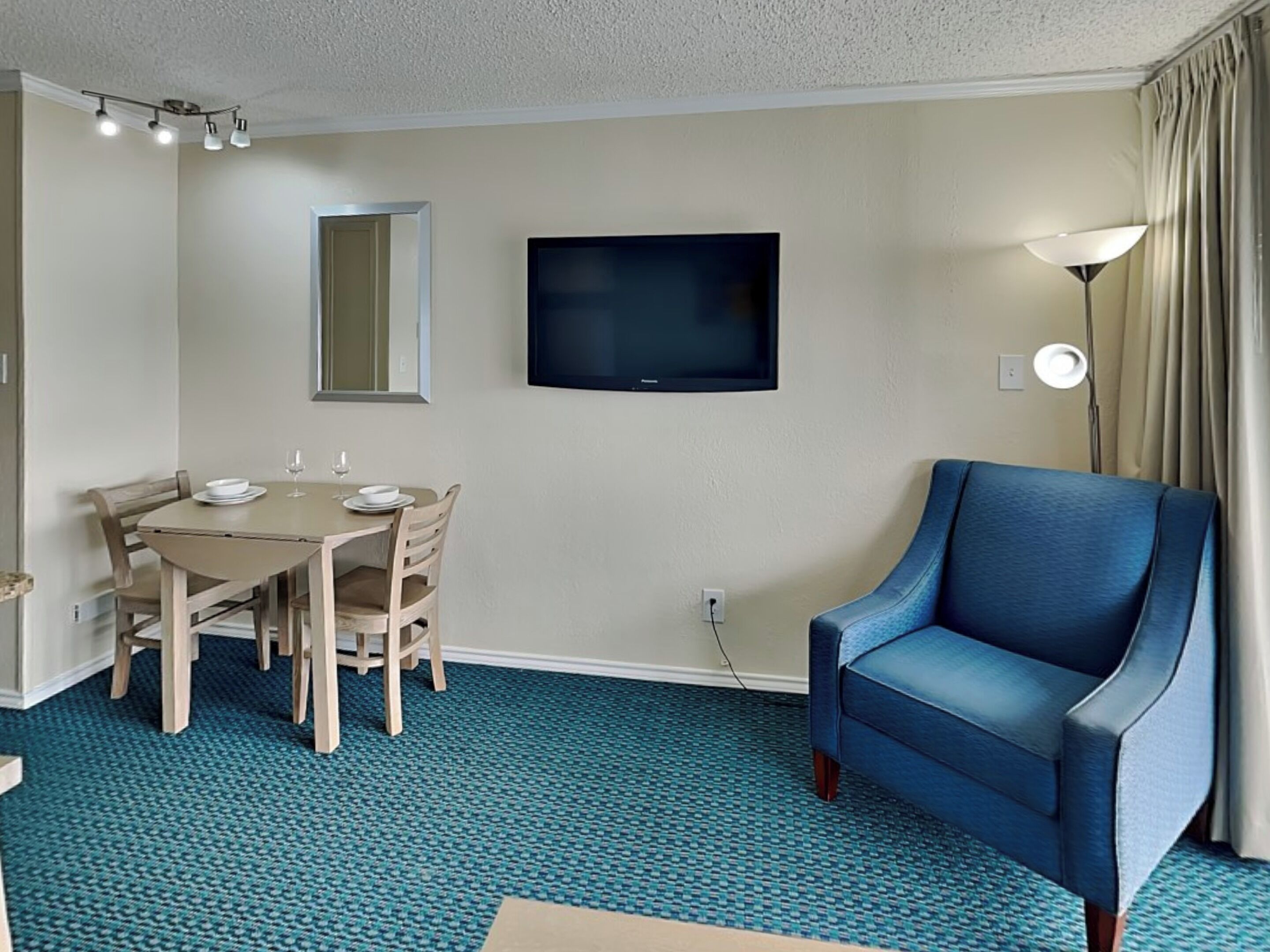 Habitación de hotel con una mesa de comedor, sillas, un televisor montado en la pared, un sillón azul y suelo alfombrado.