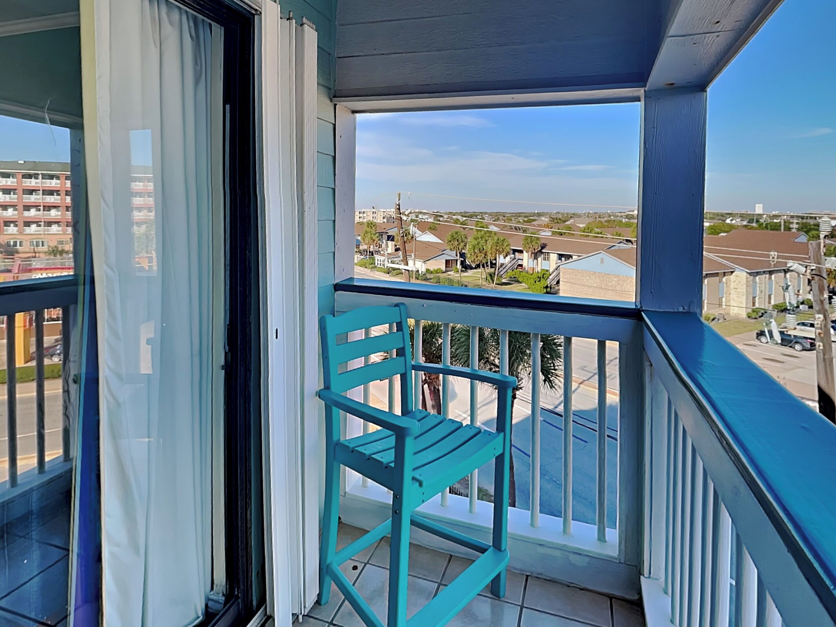 A balcony with a turquoise chair, a view of a residential area, and a building in the background.