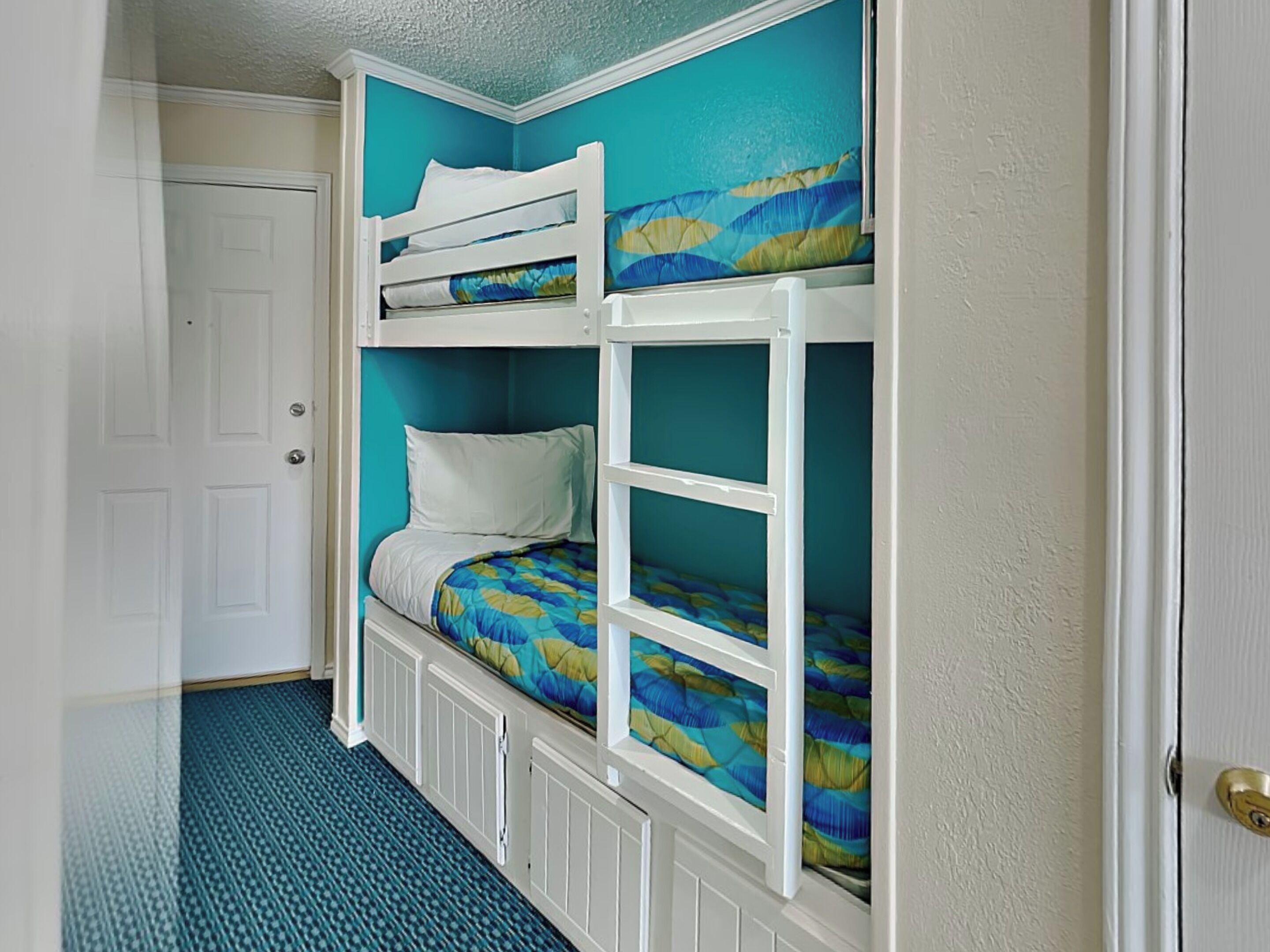 Una cama litera con dos niveles, cada uno con colchón y almohada. La habitación tiene paredes color turquesa y una puerta blanca.