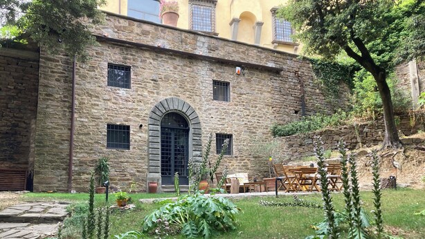 Um edifício de pedra com uma porta arqueada central, um pequeno jardim com diversas plantas e mobiliário de exterior.