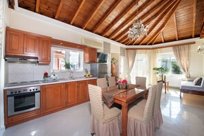 A spacious kitchen with wooden cabinets, a dining area with a table and chairs, and a living room with a sofa and a window. - Socrates Holidays Apt. 4 with Pool near the Beach (PEO Kissamou Chanion 27, Kamisiana 730 06, Greece)