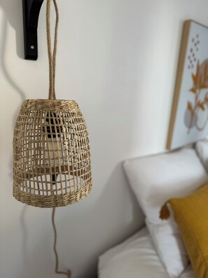 Luminária pendente tecida suspensa por um suporte preto, uma parede branca e uma cama com roupa de cama branca e um travesseiro cor de mostarda.