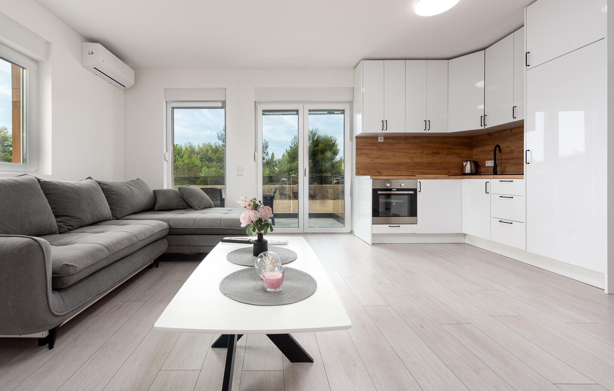 A modern kitchen with white cabinets and a wooden countertop, a grey sofa, and a dining table with a vase of flowers.