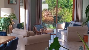 Ein Wohnzimmer mit zwei Sofas, einem Esstisch und Stühlen. Ein großes Fenster mit Vorhängen, eine Pflanze und ein Korbstuhl sind ebenfalls vorhanden.