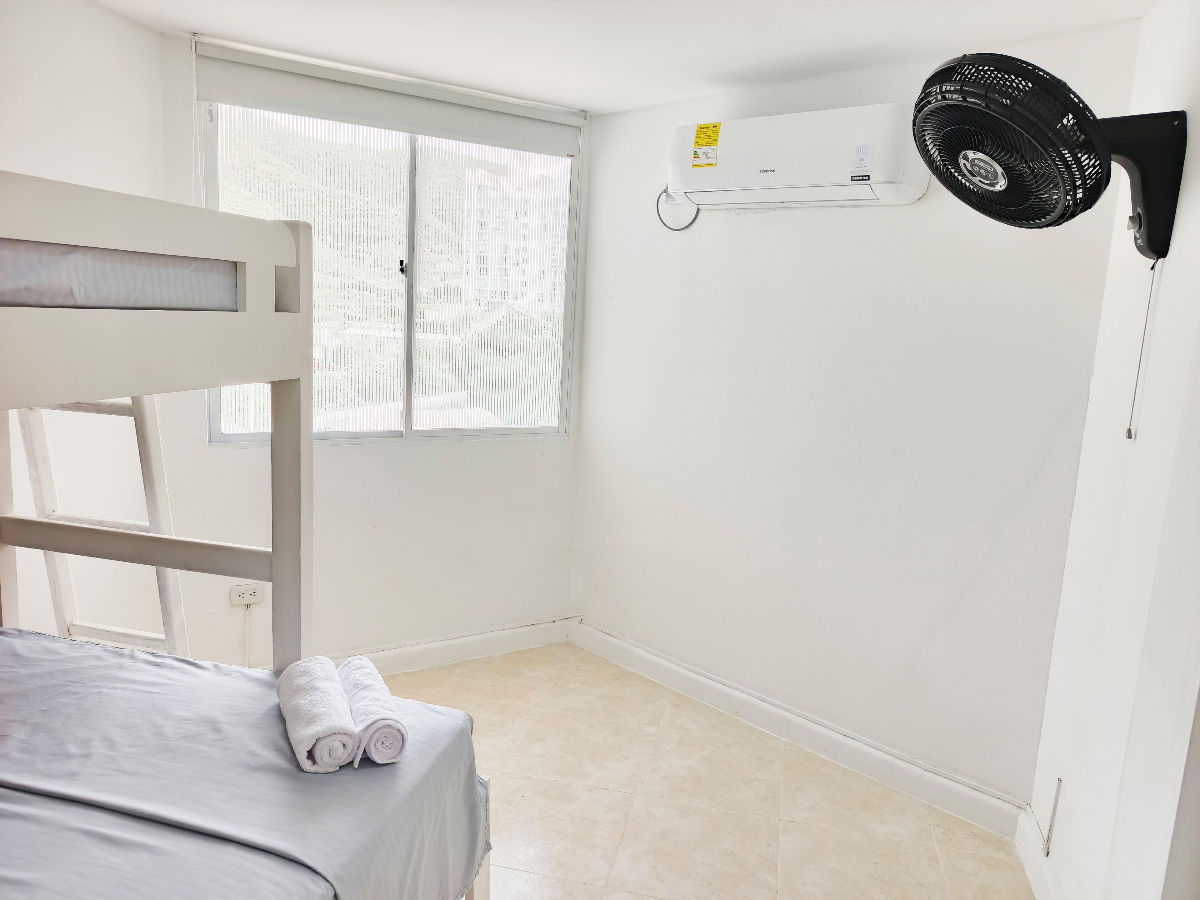 Una habitación con una cama, una ventana, aire acondicionado de pared y un ventilador de pie.