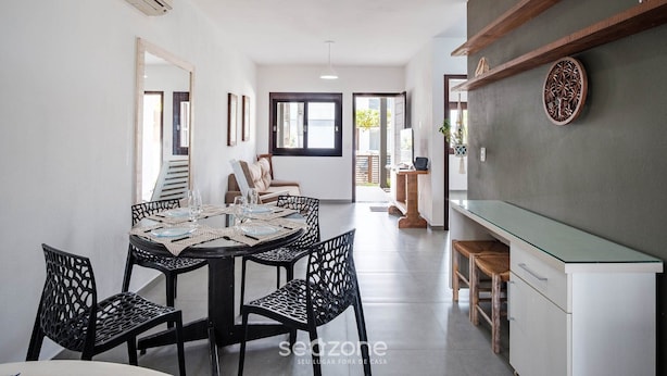 A dining area with a round table, black chairs, and a glass-top table. There is a kitchen area with a white countertop and wooden shelves. - House w/ 3 Bedrooms and Barbecue Lpb0000 (Garopaba)