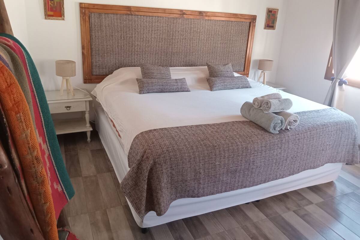 A neatly made bed with a grey headboard, white bedding, and grey pillows. There are two bedside tables with lamps, a folded towel on the bed, and a colorful blanket draped over the side.