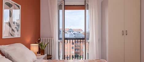 Un dormitorio con cama, mesita de noche, lámpara, planta en maceta y vista a edificios a través de una ventana.
