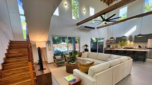 Amplia sala de estar con un sofá blanco grande, una escalera de madera y una cocina equipada con electrodomésticos modernos y taburetes de bar.