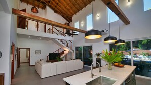 Un amplio salón con un sofá blanco, una isla de cocina con pileta y una escalera que sube a un nivel superior.