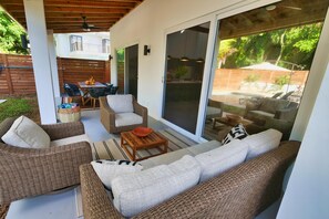 An outdoor patio with wicker furniture, a wooden table, and a glass door leading to a pool area. - Modern Comfort Just Minutes from West Bay Beach (West Bay)