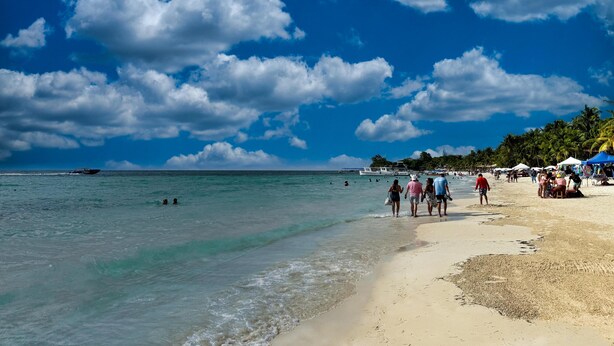 Una playa con agua turquesa cristalina, gente caminando por la orilla y un barco a lo lejos.