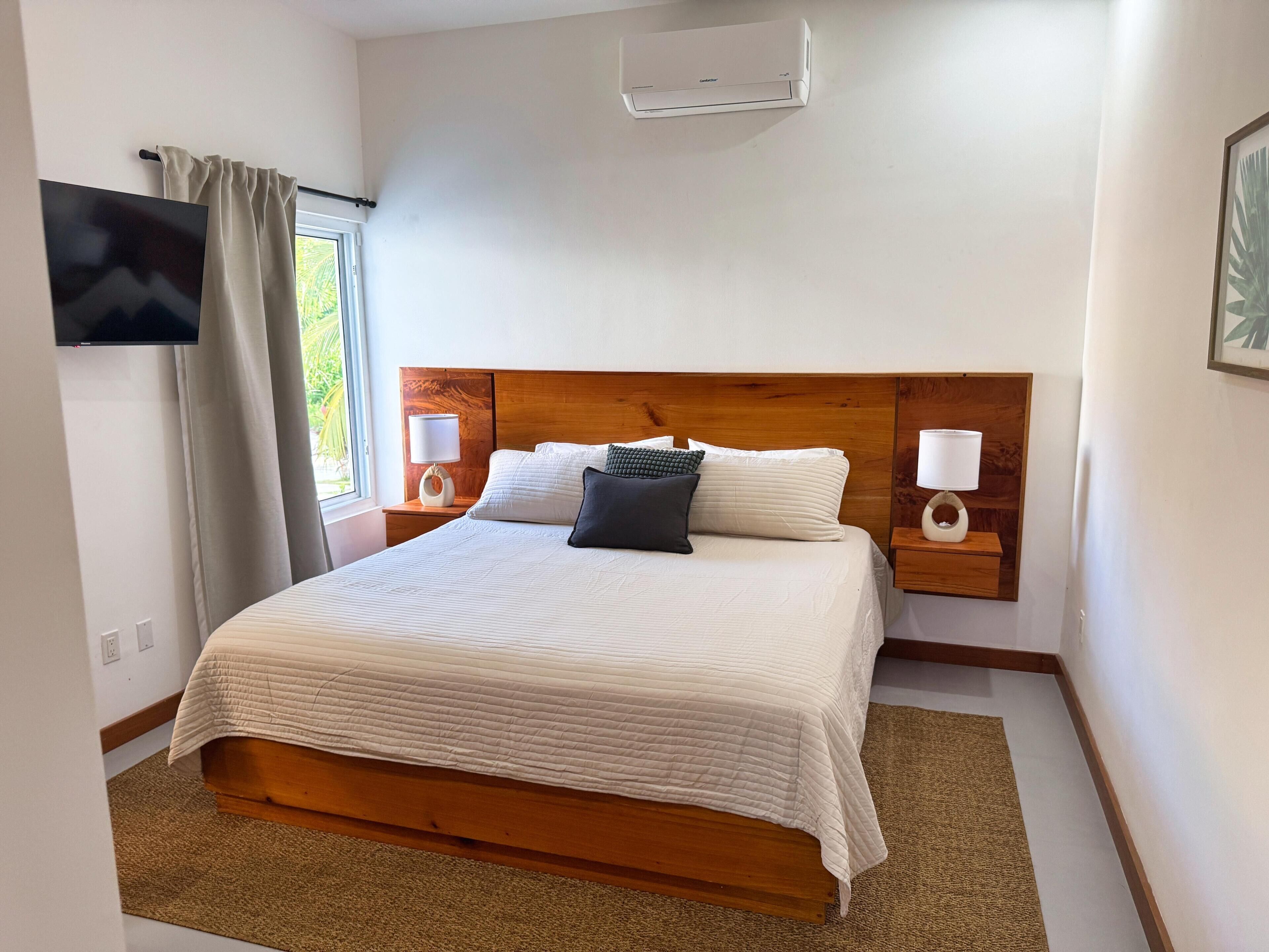 A neatly made bed with a wooden headboard, two bedside tables with lamps, a flat-screen TV mounted on the wall, and a window with curtains.