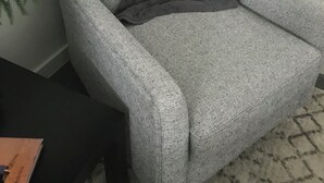 Ein grauer Sessel mit einer dunkelgrauen Decke, platziert vor einer weißen Wand mit einer Pflanze links davon.