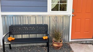 Eine schwarze Bank mit orangefarbenen Kürbissen daneben, ein Blumentopf und eine rote Tür.