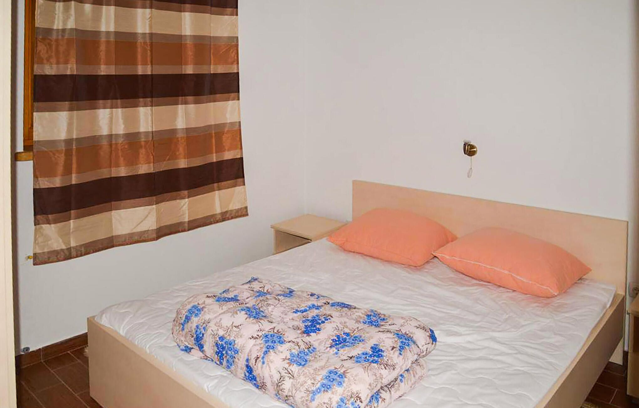 Een eenpersoonsbed met een bloemenprint sprei, twee perzikkleurige kussens, een houten hoofdbord, een nachtkastje en een gestreept gordijn.