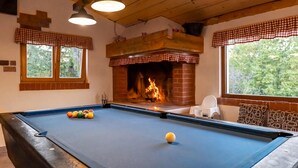 Una mesa de billar con bolas, una chimenea y ventanas con cortinas cuadriculadas.