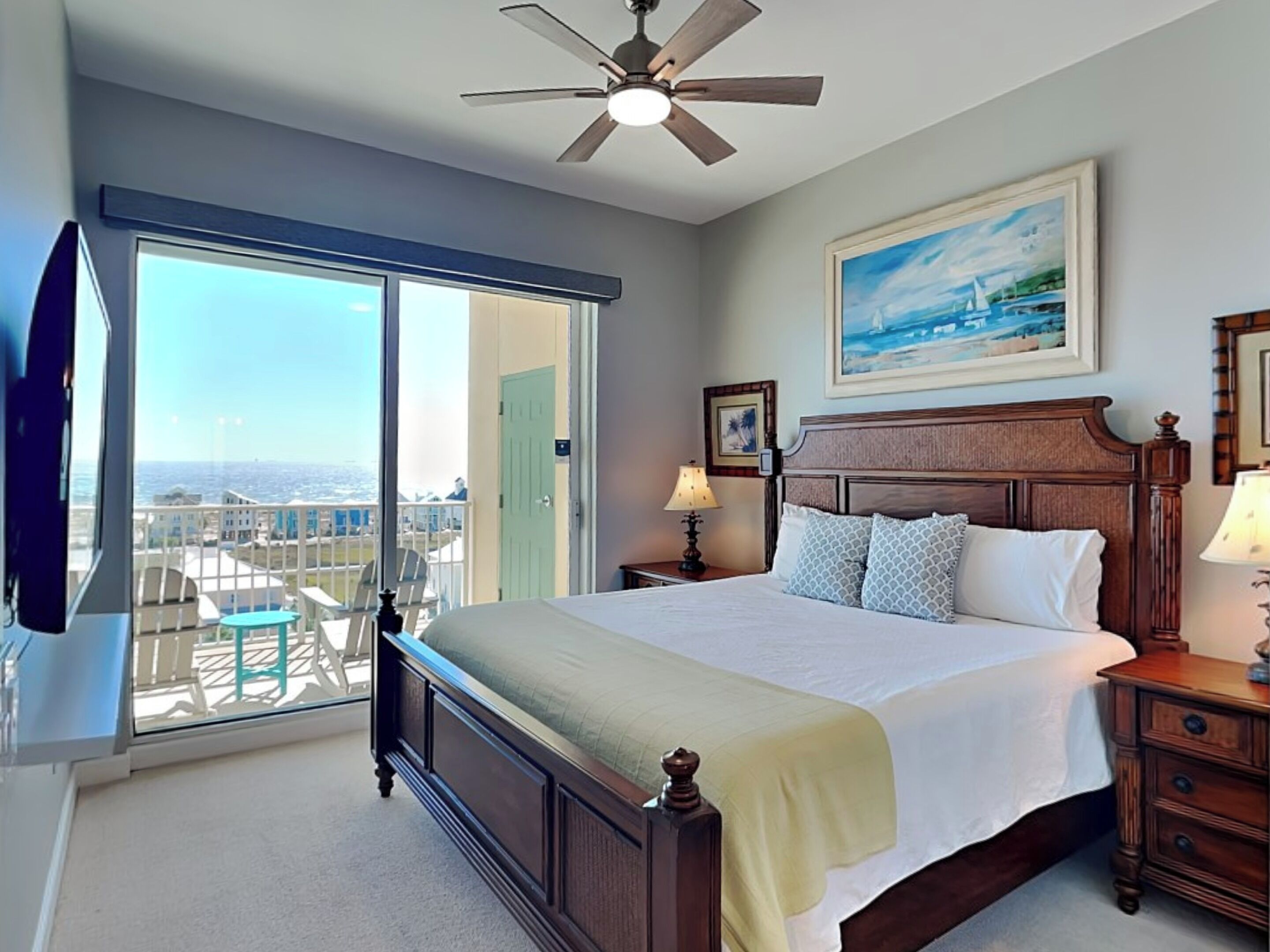 A bedroom with a large bed, a ceiling fan, a painting, and a view of the ocean through a sliding door.