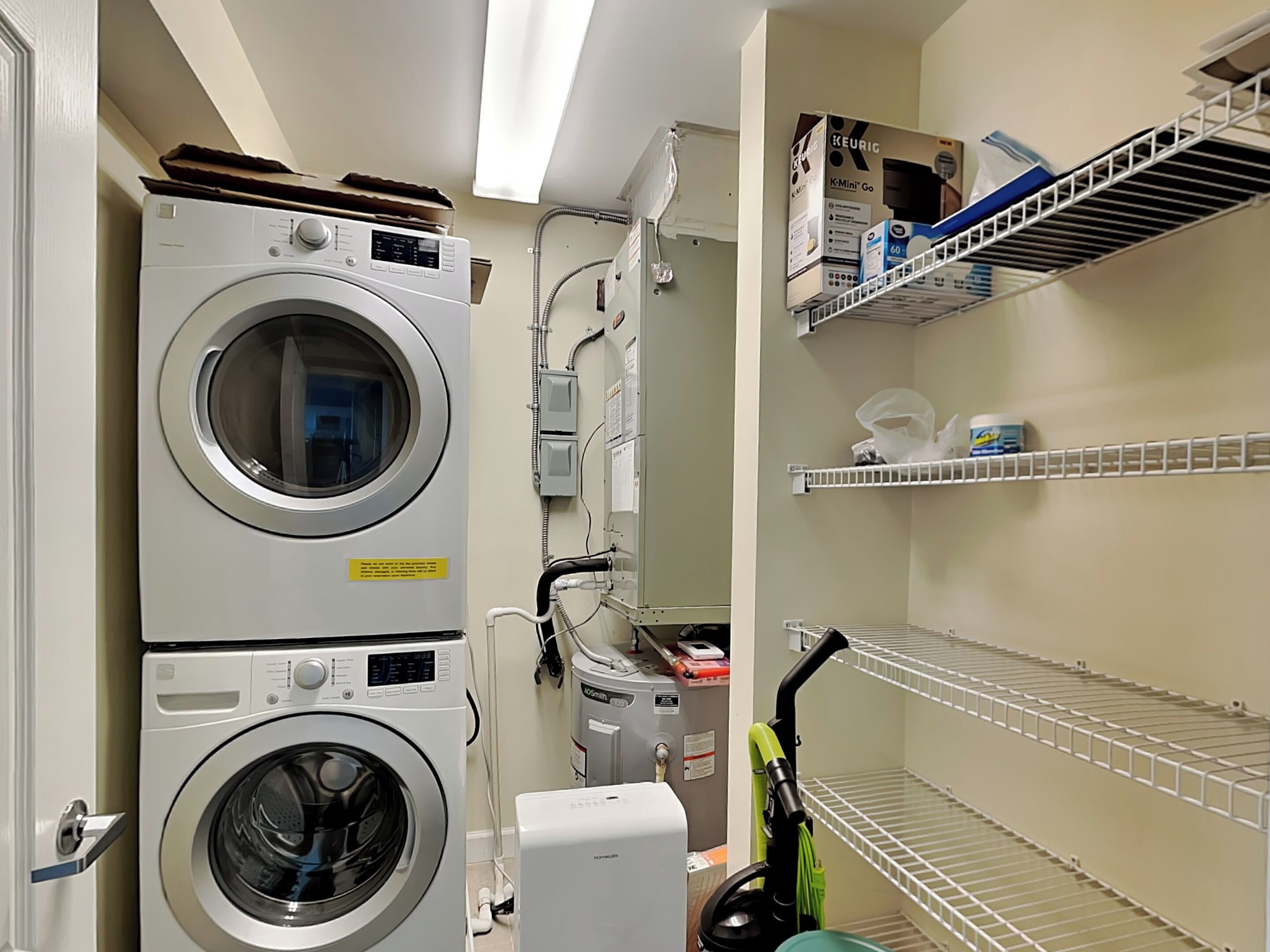 A laundry room with stacked washers and dryers, a vacuum cleaner, and shelves with cleaning supplies.