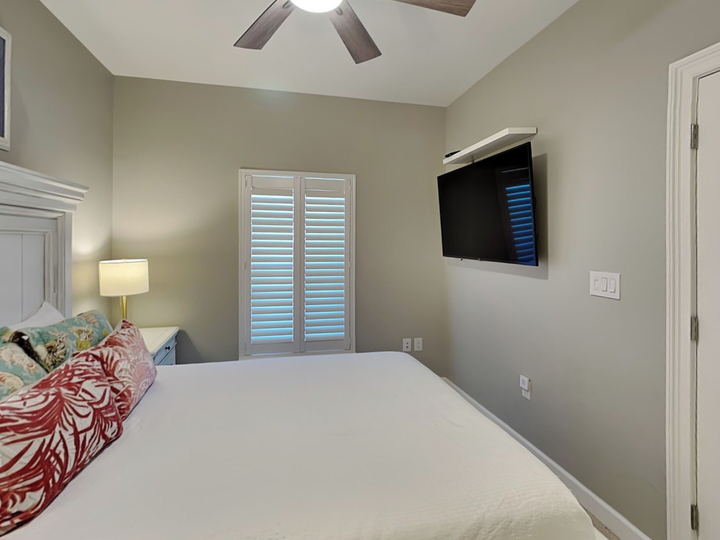 A bedroom with a bed, a ceiling fan, a TV mounted on the wall, and a window with blinds.