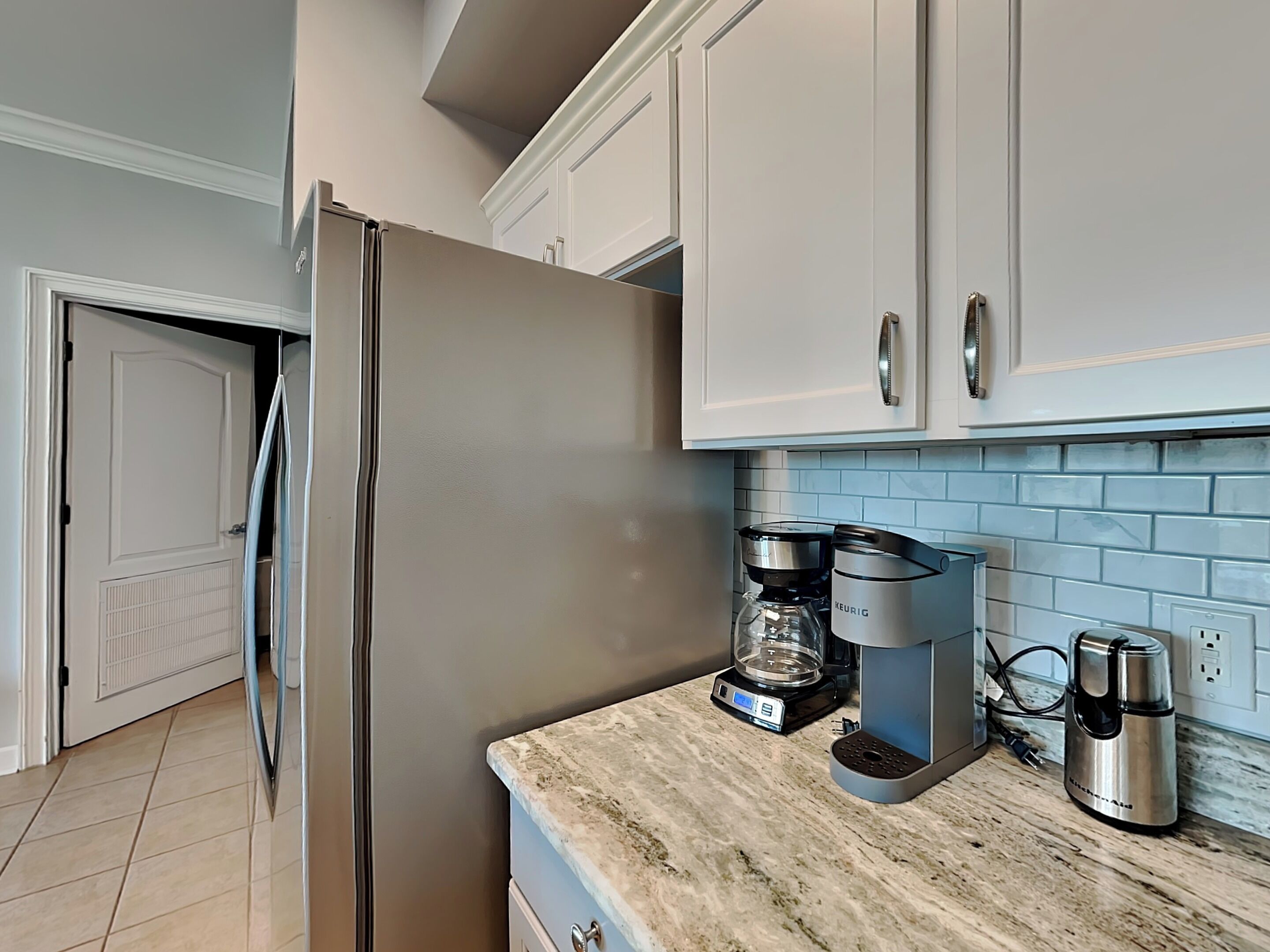A kitchen with white cabinets, a stainless steel refrigerator, a coffee maker, and a kettle on a marble countertop.