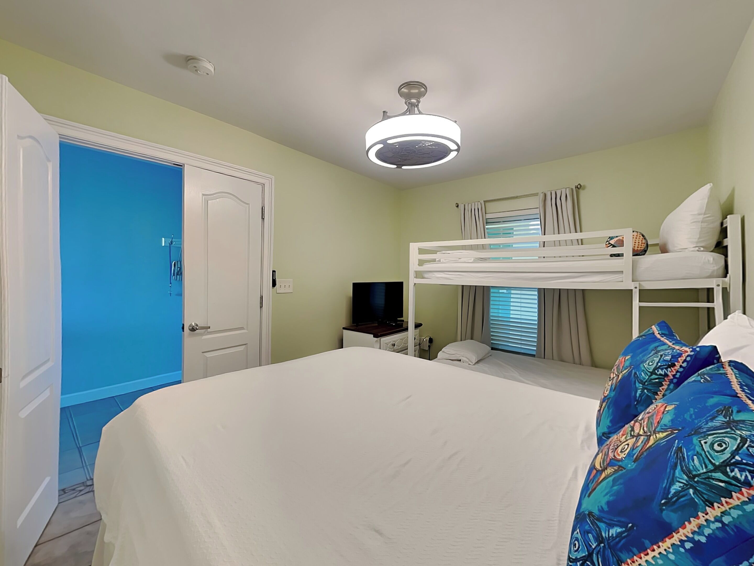 Een kamer met een stapelbed, een bureau met een computer, een televisie en een deur naar een andere kamer.