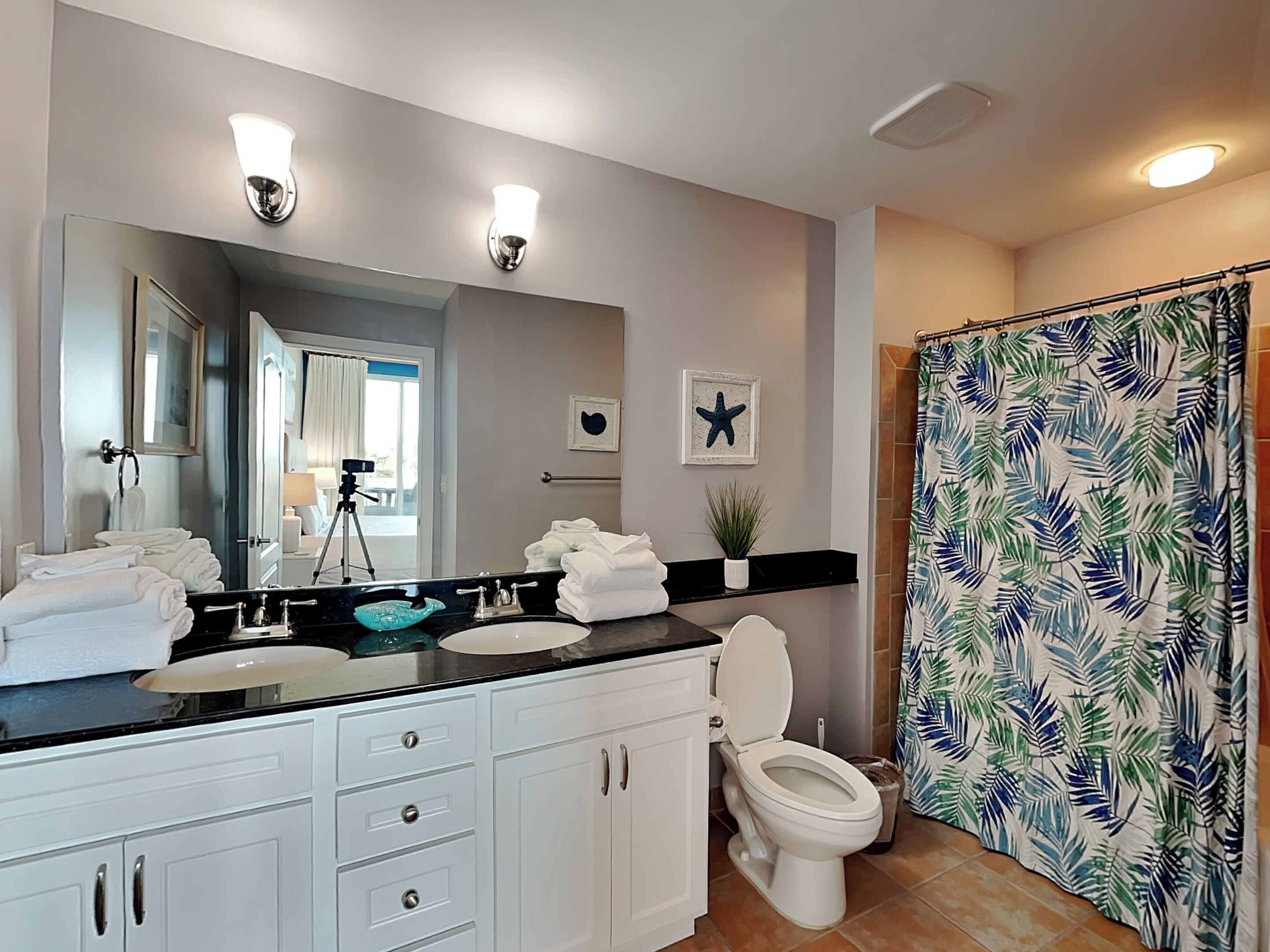 A modern bathroom with a large mirror, white cabinets, a black countertop, and a shower curtain with a tropical leaf pattern.