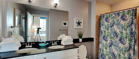 A modern bathroom with a large mirror, white cabinets, a black countertop, and a shower curtain with a tropical leaf pattern.