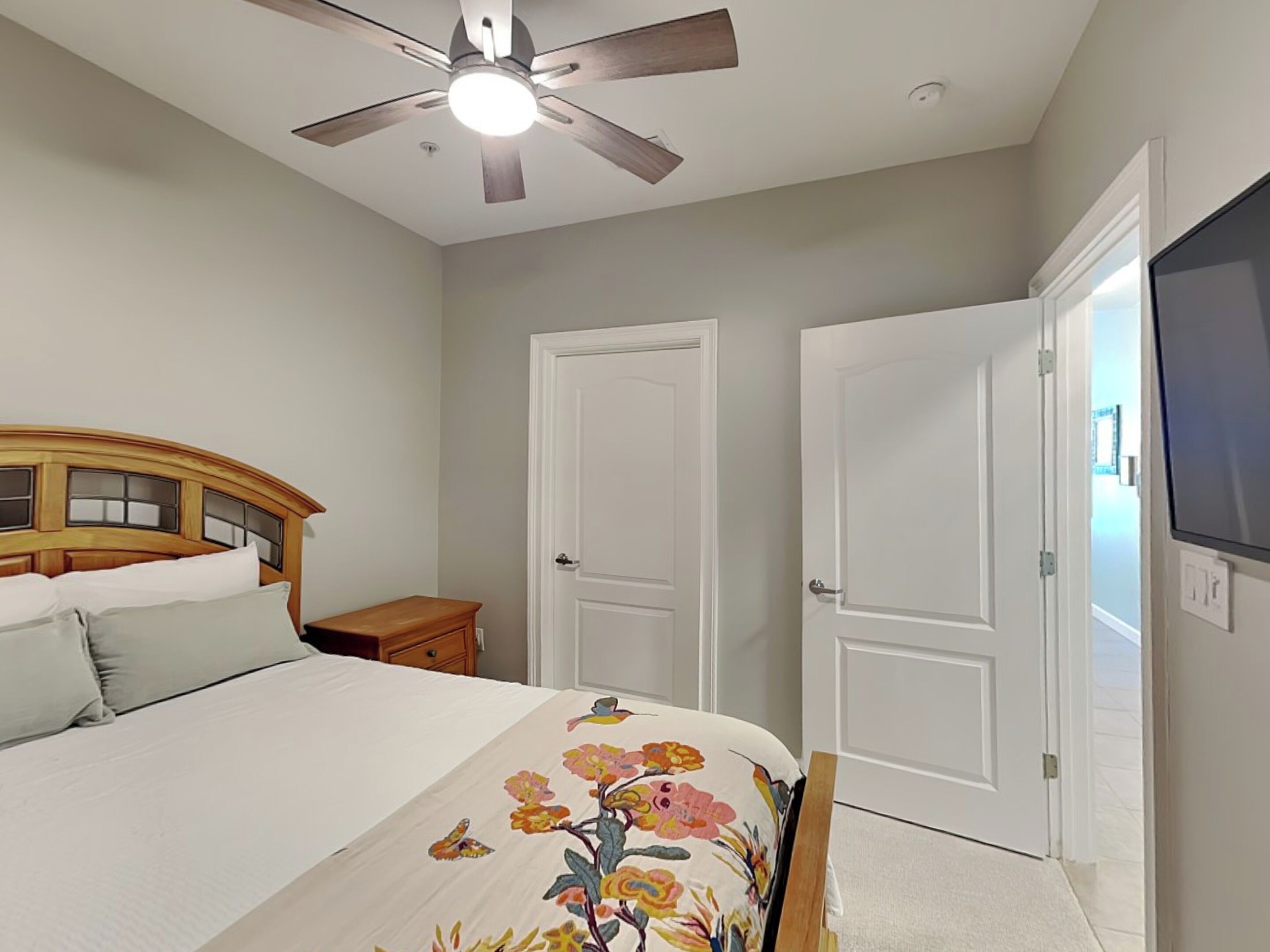 Un dormitorio con cama, ventilador de techo, dos puertas blancas y un televisor en la pared.