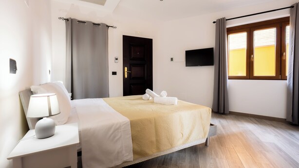 A hotel room with a bed, a nightstand with a lamp, a TV mounted on the wall, and a window with curtains. - Il Piccolo Borghetto - Guest House Stazione Tiburtina (Roma)