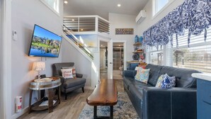 Ein modernes Wohnzimmer mit einer schwarzen Ledersofa, einem Holztisch und einem an der Wand befestigten Fernseher. Eine Treppe führt zu einer oberen Ebene, und ein großes Fenster bietet Ausblick ins Freie.