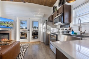 A modern kitchen with a stove, microwave, and sink, a dining area with a fire feature, and a large window. - Grace's Joy | 2 Bedrooms + Loft | Electric Fireplace (Fredericksburg)
