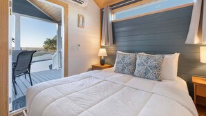 Quarto com uma cama grande, uma mesinha de cabeceira, uma luminária e vista para um deck externo por uma porta aberta.