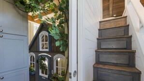 Un escalier en bois avec une rampe, une porte dotée d’une fenêtre, et une petite structure ressemblant à une maison, avec des fenêtres en arc et des plantes suspendues.