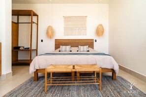 A bedroom with a wooden bed, two pillows, a woven bench, a macramé wall hanging, and a wardrobe.