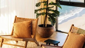 A room with a view of the ocean, featuring a large potted plant, two wicker chairs, and a small table.