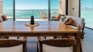 A dining area with a wooden table and chairs, a vase, and a view of the sea through large windows.