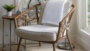 A wicker chair with a cushion, a small table with a plant, and a floor lamp in a room with a view of greenery outside.