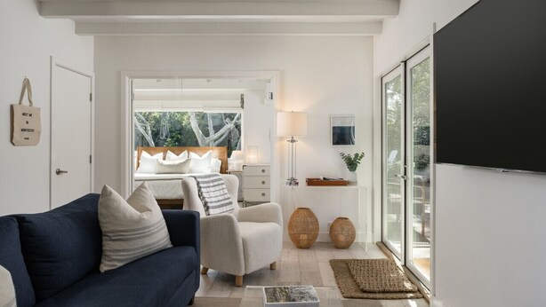 A modern living room with a sofa, armchair, and a coffee table. There is a large window with a view of trees and a door leading to a bedroom.