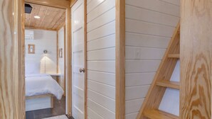 A wooden interior with a hallway leading to a bedroom, a staircase, and a visible bed. - Tiny Treasure | 1.5 Baths | Sleeps 6 | Dual Porches (Fredericksburg)