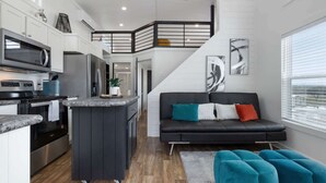 Eine moderne Küche mit einer zentralen Kochinsel, ein schwarzes Sofa mit bunten Kissen und ein Blick ins Freie durch große Fenster.