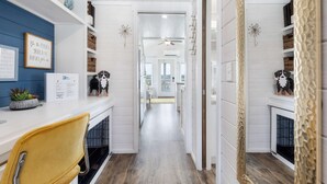 Een gang met witte muren, houten vloeren en een gele stoel. Er staan planken met manden en decoratieve objecten.