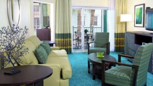 Une chambre d’hôtel avec un canapé jaune, des fauteuils verts, une petite table ronde, un téléviseur et un balcon avec vue.