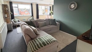 A living room with a beige sofa, a green and white checkered throw, a clock on the wall, and a view of buildings outside. - Meriden Apartment Retreat - Sleeps 7 - Near Nec (Solihull)