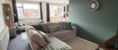 A living room with a beige sofa, a green and white checkered throw, a clock on the wall, and a view of buildings outside.