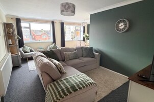 A living room with a beige sofa, a green and white checkered throw, a clock on the wall, and a view of buildings outside.