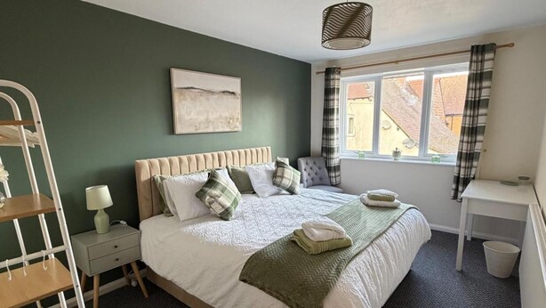 A bedroom with a bed, a bedside table, a lamp, a window with curtains, and a view of buildings outside. - Meriden Apartment Retreat - Sleeps 7 - Near Nec (Solihull)
